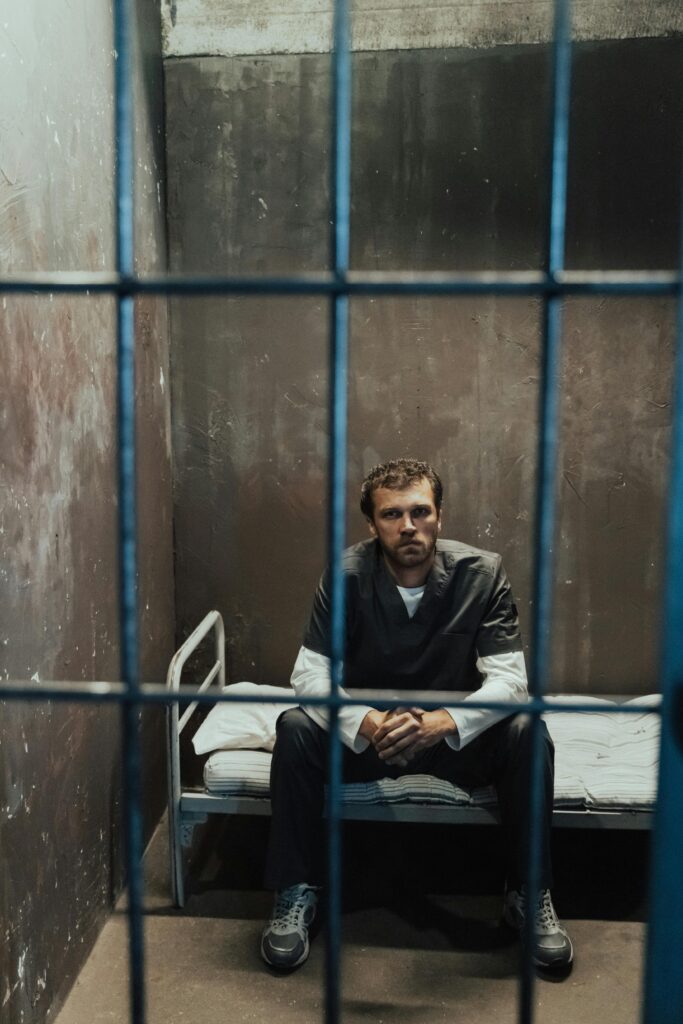 A male prisoner in uniform sits on a bed in a dimly lit jail cell, viewed through bars.