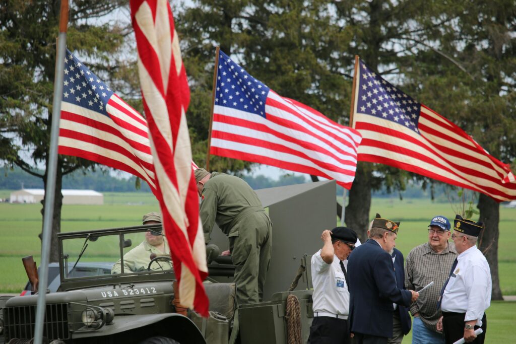 Group of veterans and soldiers by military vehicle with American flags in the background, symbolizing patriotism.